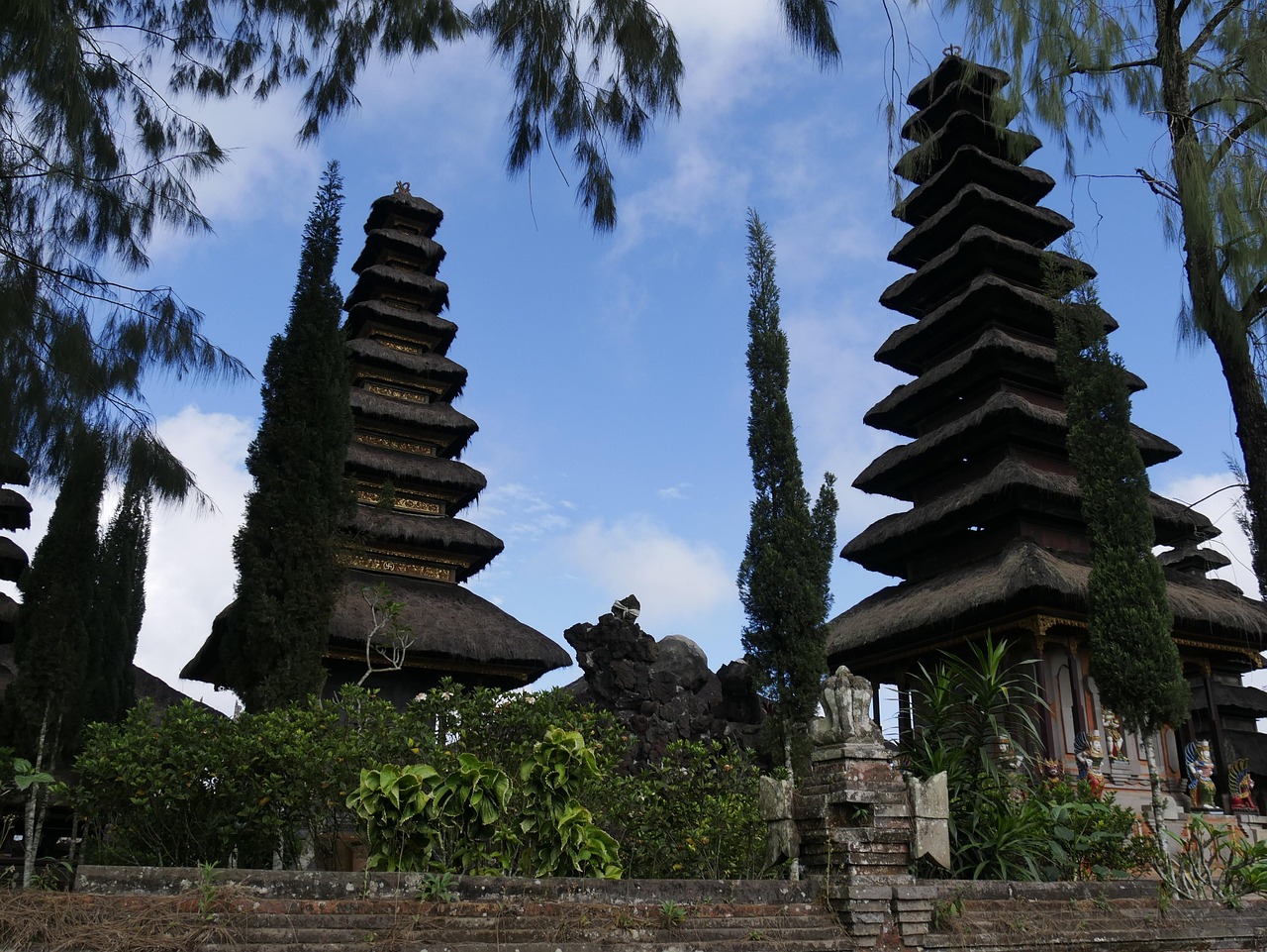 Traditional Balinese temple towers rising above lush greenery and stone shrines under a soft blue sky, reflecting the island&rsquo;s spiritual landscape beyond its busy tourist areas.